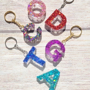 May include: Six colorful glitter resin letter keychains, each with a silver or gold key ring. The letters are A, C, D, G, T, and O. The letters are all different colors, including pink, red, purple, blue, and teal.
