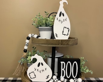 May include: A two-tiered wooden tray with a black and white checkered tablecloth. The tray is decorated with black and white ghost ornaments, a black and white beaded garland, and a black wooden block with the word "BOO" in white letters. The tray is also decorated with greenery and a small white pot.