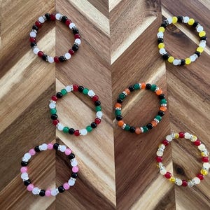 May include: Six colorful beaded bracelets with different color combinations, including black, white, red, green, orange, and yellow beads. The bracelets are arranged on a wooden surface.