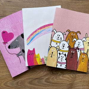 May include: Three pink cards with illustrations. The first card features a grey dog holding a pink heart-shaped balloon. The second card has a rainbow. The third card has a group of dogs with different coloured fur.