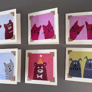 May include: Six handmade greeting cards with quirky animal illustrations. Each card showcases a different design, including cats, bears, and dogs, framed within a square with decorative corner mounts. The cards are set against a neutral backdrop.