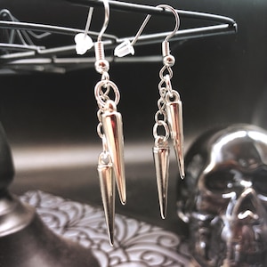 May include: Silver-colored dangle earrings with a spike design. Each earring features a chain with three cone-shaped spikes. The earrings hang from a silver hook. The background includes a decorative metal object and a skull.