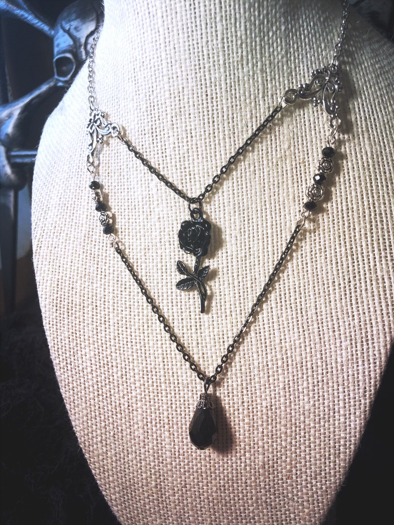 May include: A silver chain necklace with a black rose pendant and a black teardrop pendant. The necklace is layered with black beads and a silver filigree charm.