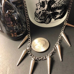 Pu&ograve; includere: Una collana a catena d'argento con sei punte d'argento appese. La collana &egrave; appoggiata su una superficie nera con una moneta d'argento accanto.
