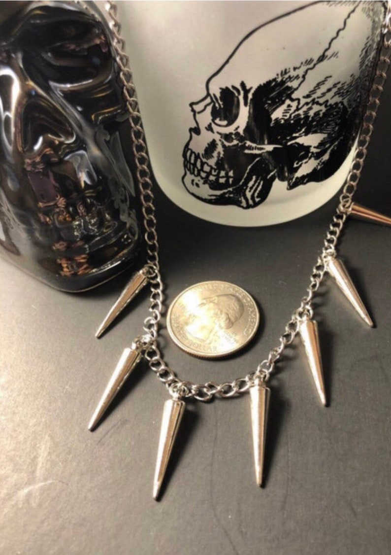 Pu&ograve; includere: Una collana a catena d'argento con sei punte d'argento appese. La collana &egrave; appoggiata su una superficie nera con una moneta d'argento accanto.