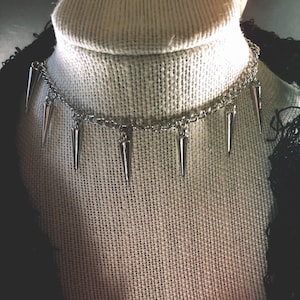 Pu&ograve; includere: Un collare choker argentato con pi&ugrave; punte appuntite che pendono dalla catena.