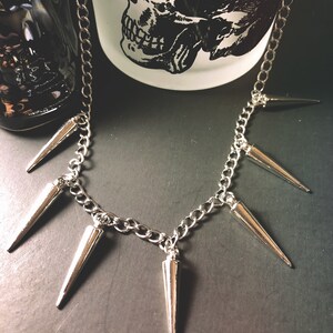 Pu&ograve; includere: Una collana a catena d'argento con pi&ugrave; punte d'argento appese. La collana &egrave; su una superficie grigio scuro.