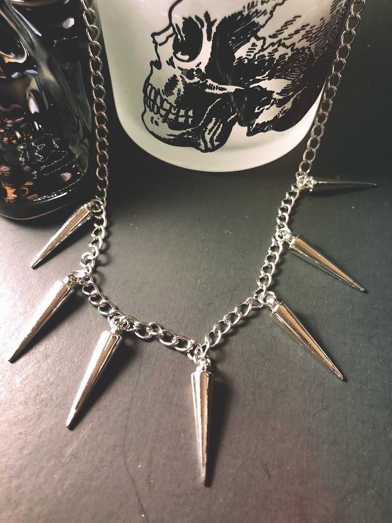 Pu&ograve; includere: Una collana a catena d'argento con pi&ugrave; punte d'argento appese. La collana &egrave; su una superficie grigio scuro.