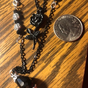 May include: A black chain necklace with a silver pendant and black rose charms. The necklace also features black beads and a teardrop-shaped black crystal.
