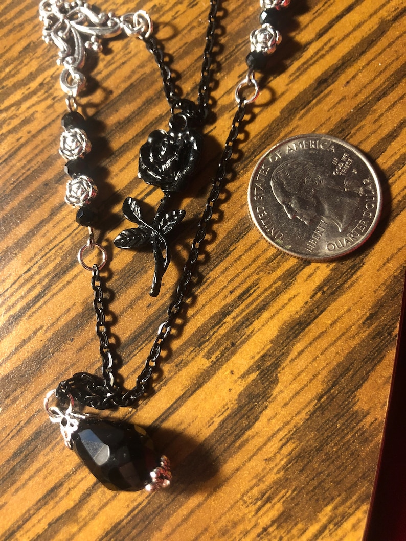 May include: A black chain necklace with a silver pendant and black rose charms. The necklace also features black beads and a teardrop-shaped black crystal.