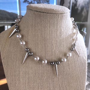 May include: A silver-toned necklace with faux pearls and spiked accents. The necklace is displayed on a beige mannequin. The necklace features alternating faux pearls and silver-toned beads, with several silver spikes.