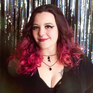 May include: A woman with pink and dark hair wearing a black v-neck top with a black choker necklace with a rose pendant. She is standing in front of a silver and black sparkly background.