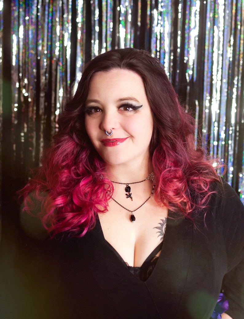 May include: A woman with pink and dark hair wearing a black v-neck top with a black choker necklace with a rose pendant. She is standing in front of a silver and black sparkly background.