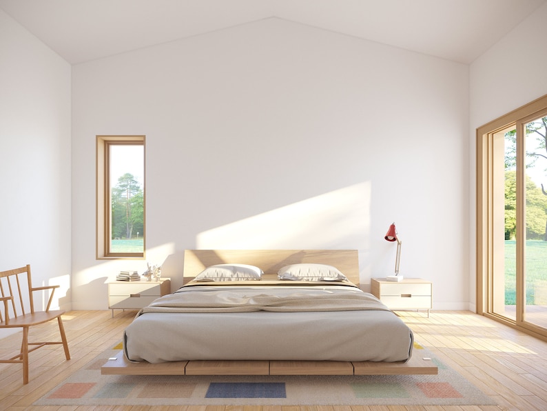 May include: A minimalist bedroom with a light wood bed frame, two bedside tables, and a wooden chair. The bed is covered with a beige comforter and pillows. A red desk lamp sits on one of the bedside tables. A large window and a smaller window provide natural light.