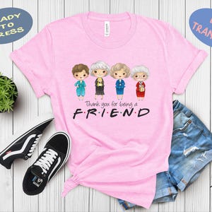 May include: Pink t-shirt featuring a cartoon illustration of four women in colorful outfits. The text "Thank you for being a F.R.I.E.N.D" is printed below the characters. Black and white sneakers and denim shorts are also in the image.