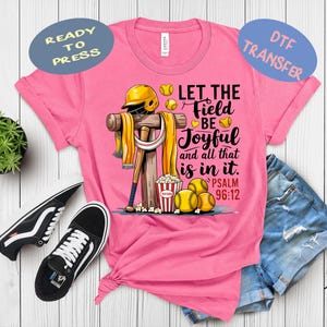 May include: Pink t-shirt with a baseball-themed graphic. The design includes a baseball bat, helmet, and baseballs, with the text "Let the Field Be Joyful and all that is in it. Psalm 96:12". Includes a pair of black and white sneakers and denim shorts.