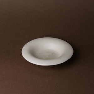 Może przedstawiać: Mała, kremowobiała ceramiczna miska z płytkim, zaokrąglonym wnętrzem. Miska ma prosty, minimalistyczny design i jest umieszczona na brązowym tle. Średnica miski wynosi około 15 cm.