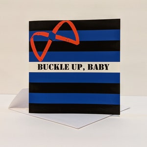 May include: A greeting card with a black and blue striped background. A red buckle is in the top left corner. The text "BUCKLE UP, BABY" is in white letters on a white stripe.
