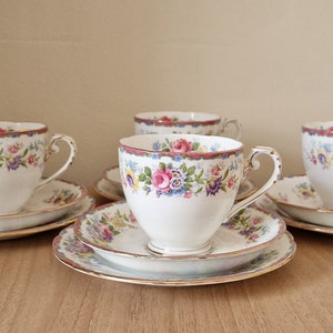 Puede incluir: Juego de cuatro tazas de té y platillos florales. Cada pieza presenta un delicado patrón floral en tonos de rosa, azul y amarillo, con un borde dorado. Las tazas y los platillos son de porcelana blanca.