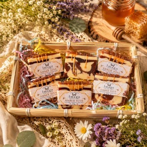 May include: A rectangular woven basket containing several handcrafted "Imago Amber Hive" soap bars. The soaps have a layered design with amber-coloured accents. The image also features honey, a honeycomb, and flowers, suggesting a natural theme.