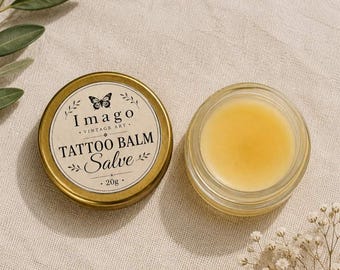 Op de afbeelding: Twee containers met tatoeagebalsem. Eén is een kleine, ronde, goudkleurige blik met de woorden "Imago Vintage Art Tattoo Balm Salve" en een vlinderillustratie. De andere is een kleine, heldere glazen pot gevuld met een gele balsem. De balsem is 20g.