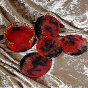 May include: Set of four red and black resin coasters with gold flecks. The coasters are round and have a marbled effect. One coaster is a larger square shape with a lip around the edge.