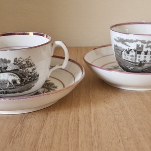 May include: Two vintage teacups and saucers with black and white pastoral scenes. One cup features a scene with sheep, the other features a scene with a house and trees. Both cups have a gold rim and a pink rim on the saucer.