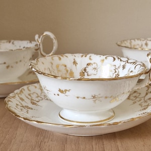 Puede incluir: Tres tazas de té blancas con ribetes dorados y diseños florales. Las tazas y los platillos están dispuestos sobre una superficie de madera.