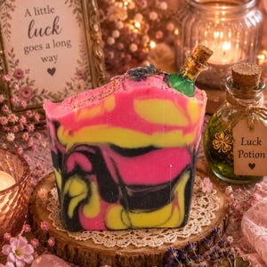 May include: A vibrant soap bar with pink, yellow, and black marbling, adorned with a miniature bottle. The soap rests on a wooden slice with a lace doily. A framed sign reads "A little luck goes a long way."
