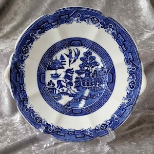 May include: A blue and white porcelain plate with a scalloped edge and a traditional Chinese willow pattern. The plate has a raised rim and a center design featuring a pagoda, a bridge, and a willow tree.