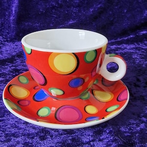 May include: A red and white ceramic teacup and saucer set with a colourful polka dot pattern. The cup has a white interior and a rounded handle. The saucer has a matching pattern.