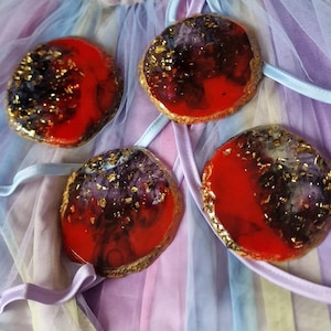 May include: Four round resin coasters with a red, black, and gold abstract design. The coasters are on a purple and blue iridescent fabric.