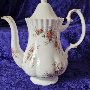 Könnte beinhalten: Eine weiße Porzellan-Teekanne mit einem floralen Muster aus rosa Rosen und grünen Blättern. Die Teekanne hat einen goldenen Rand um den Ausguss und den Henkel.