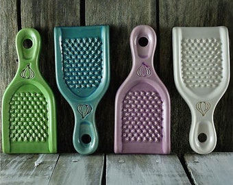 Puede incluir: Cuatro ralladores de ajo de cerámica en diferentes colores: verde, azul, morado y blanco. Cada rallador tiene un diseño de ajo y un mango.