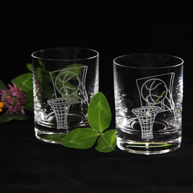 May include: Two clear, round drinking glasses with etched basketball hoop and net designs. The glasses are on a black surface, with green leaves and purple flowers in the background. The glasses are likely for whiskey or other spirits.