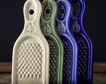 Puede incluir: Cuatro ralladores de ajo de cerámica en fila, cada uno de un color diferente: blanco roto, verde, azul y azul oscuro. Cada rallador tiene un mango con un agujero y un diseño de bulbo de ajo. La superficie para rallar está cubierta de pequeñas protuberancias.