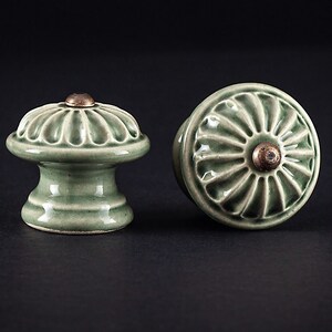 May include: Two green ceramic cabinet knobs with a raised, circular design. The knobs have a brass accent in the center.