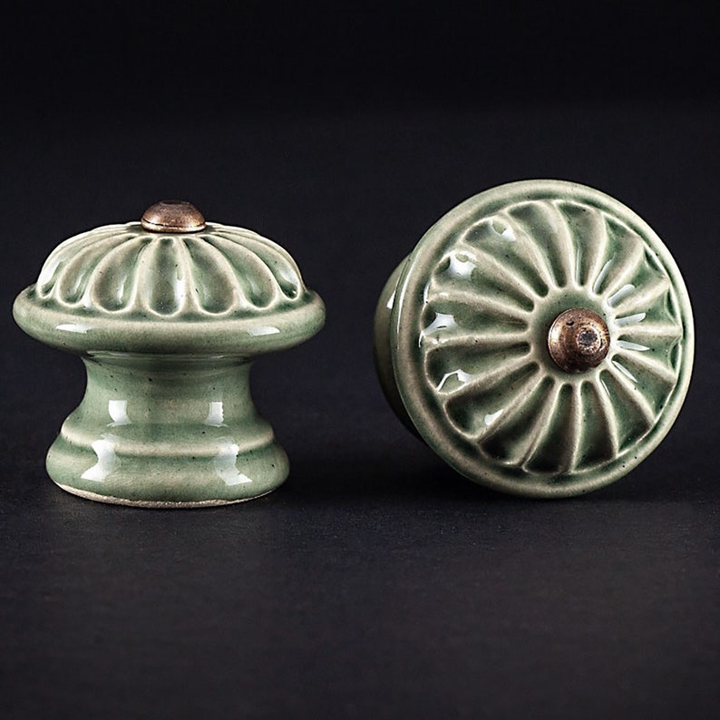 May include: Two green ceramic cabinet knobs with a raised, circular design. The knobs have a brass accent in the center.