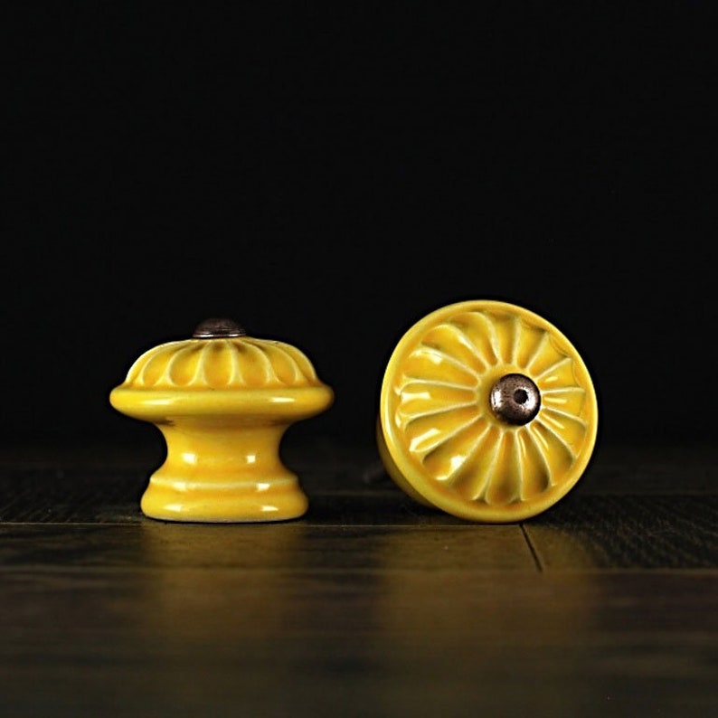 May include: Two yellow ceramic cabinet knobs with a raised, fluted design. The knobs have a round base and a raised center with a small, round, copper-colored accent.