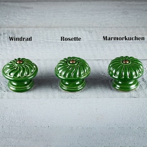 Könnte beinhalten: Drei grüne Keramik-Schrankknöpfe mit bronzefarbenen Zentren. Die Knöpfe sind mit "Windrad", "Rosette" und "Marmorkuchen" beschriftet. Die Knöpfe haben eine glänzende Oberfläche und sind auf einer hellgrauen Holzoberfläche angeordnet.