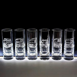 May include: Six clear glass shot glasses with etched designs of semi-trucks. The glasses are lined up in a row on a white surface.