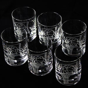 May include: Set of six clear glass tumblers with a basketball and hoop design etched on the front. The glasses are cylindrical with a thick base and are arranged on a black surface.