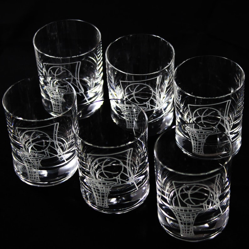 May include: Set of six clear glass tumblers with a basketball and hoop design etched on the front. The glasses are cylindrical with a thick base and are arranged on a black surface.