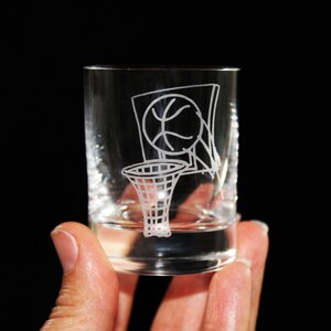 May include: A clear shot glass with a basketball and hoop design etched on the front. The design is white and features a basketball going into the net. The glass is held against a black background.