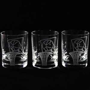 May include: Three clear, lowball glasses with etched basketball hoop and ball designs. The glasses are cylindrical with a thick base, perfect for serving cocktails or other beverages. The basketball theme is visible on each glass.