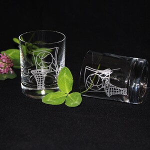 May include: Two clear, etched glass tumblers with a basketball and hoop design. One tumbler is upright, the other is tilted. Green clover and a pink flower are in the background. The glasses are on a black surface.