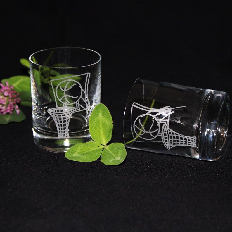 May include: Two clear, etched glass tumblers with a basketball and hoop design. One tumbler is upright, the other is tilted. Green clover and a pink flower are in the background. The glasses are on a black surface.