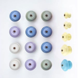 May include: A collection of ceramic cabinet knobs in various pastel colors, including blue, lavender, green, and white. Each knob has a brass-colored center. Additional knobs in a light yellow and blue are also displayed.