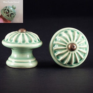 May include: Two ceramic cabinet knobs in a light green color with a raised, textured design. One knob has a round shape with a raised center, while the other has a round top with a raised, cylindrical base. Both knobs have a brass colored center.