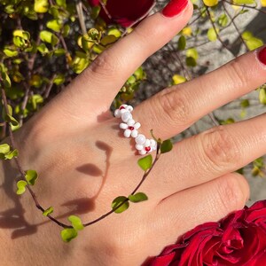 May include: A white beaded ring with small white flowers and red centers. The ring is worn on a hand with red nail polish and is surrounded by red roses.